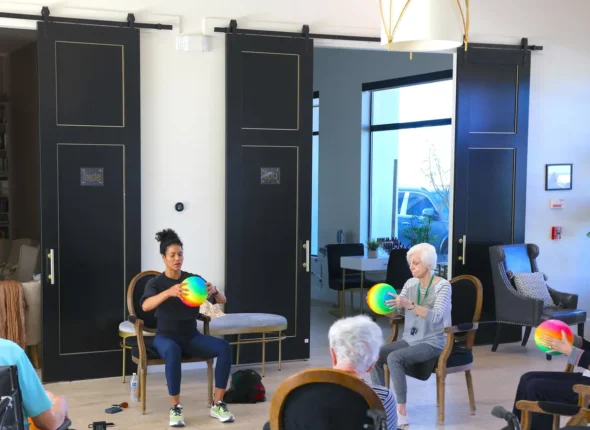 Seniors participate in a seated exercise class with colorful balls in assisted living sports room.